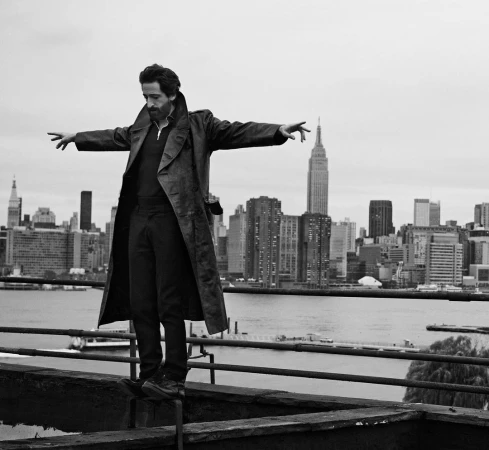Black and white shot of a man in a trench coat standing on a rooftop ledge with arms outstretched, set against the New York City skyline.
