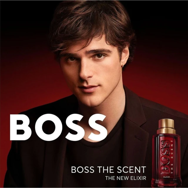 Portrait of an adult man in a dark brown blazer, posing near a bottle of Hugo Boss The Scent Elixir perfume against a dark background.