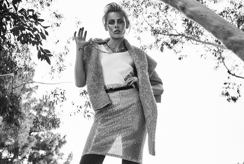 Model in knit cardigan and patterned skirt posing outdoors under tree branches in a black and white photograph by Matthew Brookes.