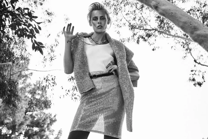 Model in knit cardigan and patterned skirt posing outdoors under tree branches in a black and white photograph by Matthew Brookes.