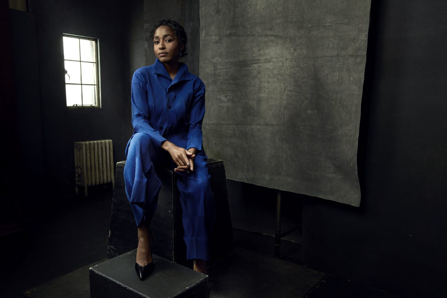 Black woman wearing a pleated blue pantsuit seated on dark boxes in a studio with a grey backdrop