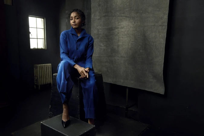 Black woman wearing a pleated blue pantsuit seated on dark boxes in a studio with a grey backdrop