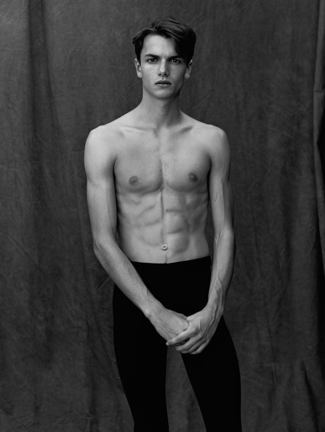 Striking black and white portrait of a shirtless adult male with dark hair, dark leggings, and folded hands, against a draped fabric background.