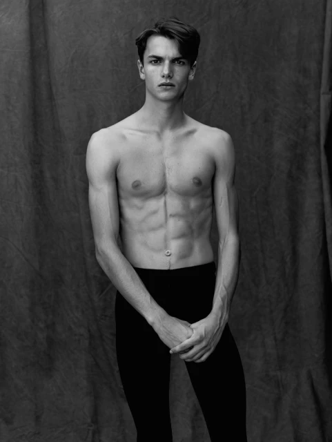 Striking black and white portrait of a shirtless adult male with dark hair, dark leggings, and folded hands, against a draped fabric background.