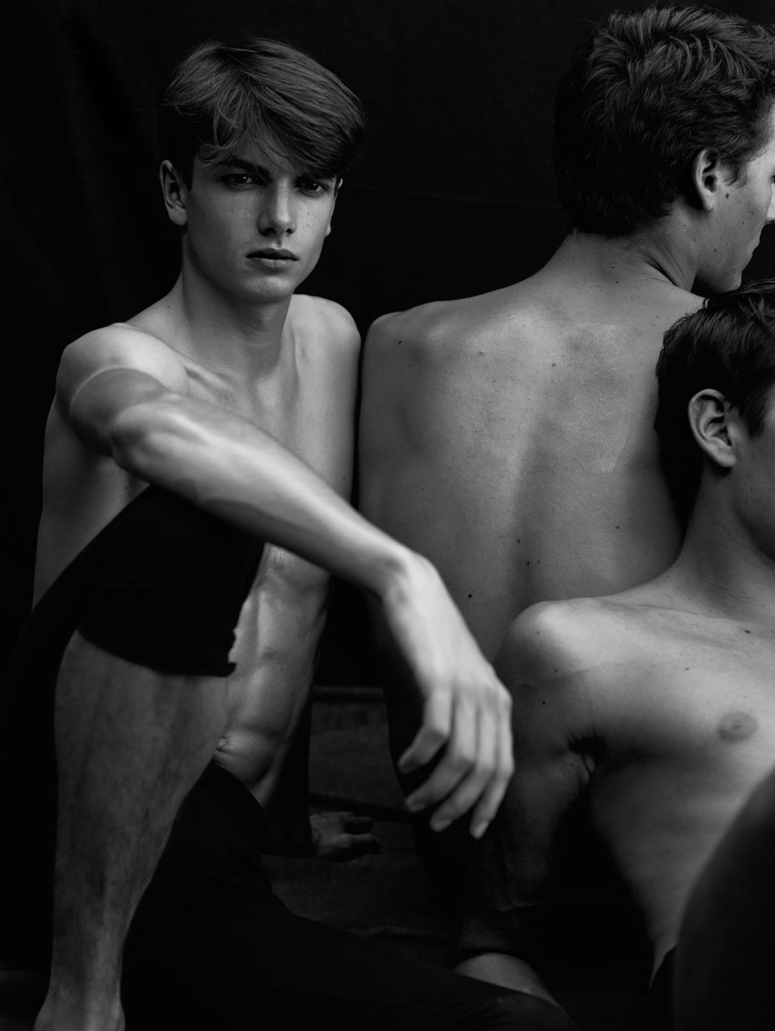 Black and white portrait of three young adult men with dark hair posing in a studio setting against a dark backdrop, artistic.