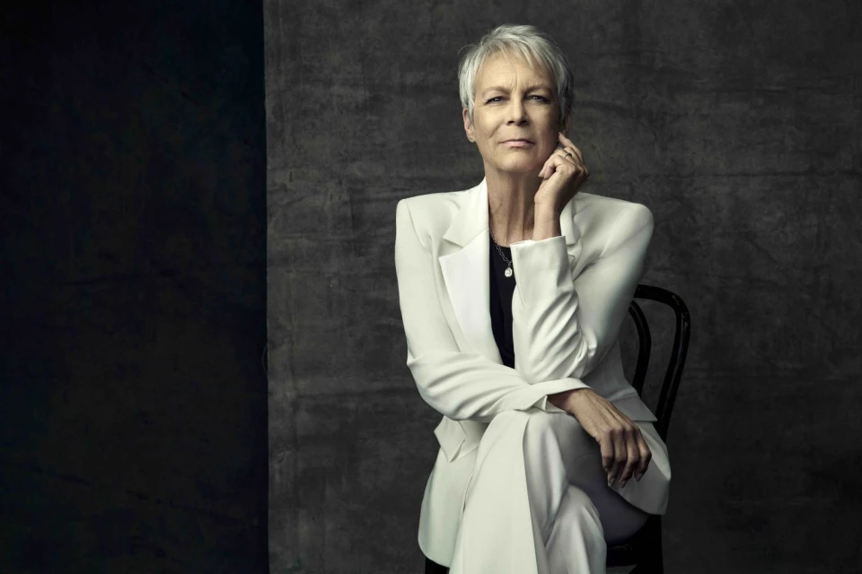 Studio portrait of Jamie Lee Curtis wearing a white suit and seated on a black chair in front of a textured grey backdrop.