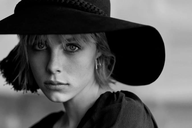 Black-and-white portrait of a young woman with short blonde hair and a wide-brimmed hat looking directly at the camera with a neutral expression.