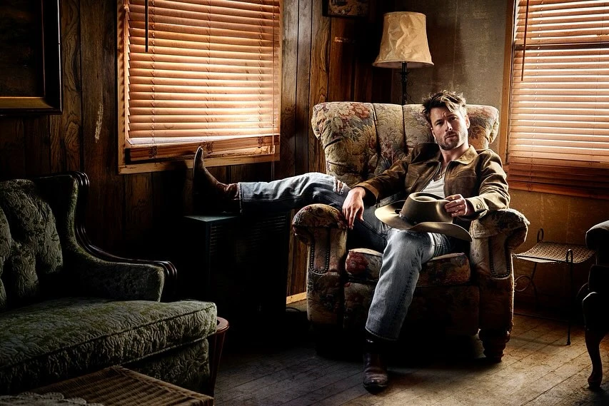 Man relaxing in an armchair in a rustic room, wearing jeans, boots, and a jacket while holding a cowboy hat, with blinds in the background.