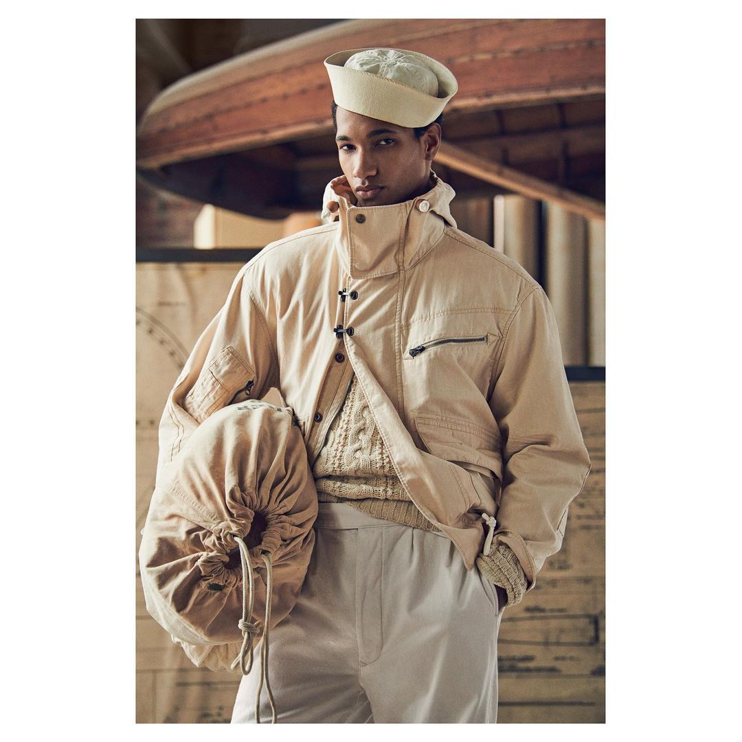 Portrait of a man wearing a sailor hat and beige jacket, holding a drawstring bag, with a vintage boat in the background.