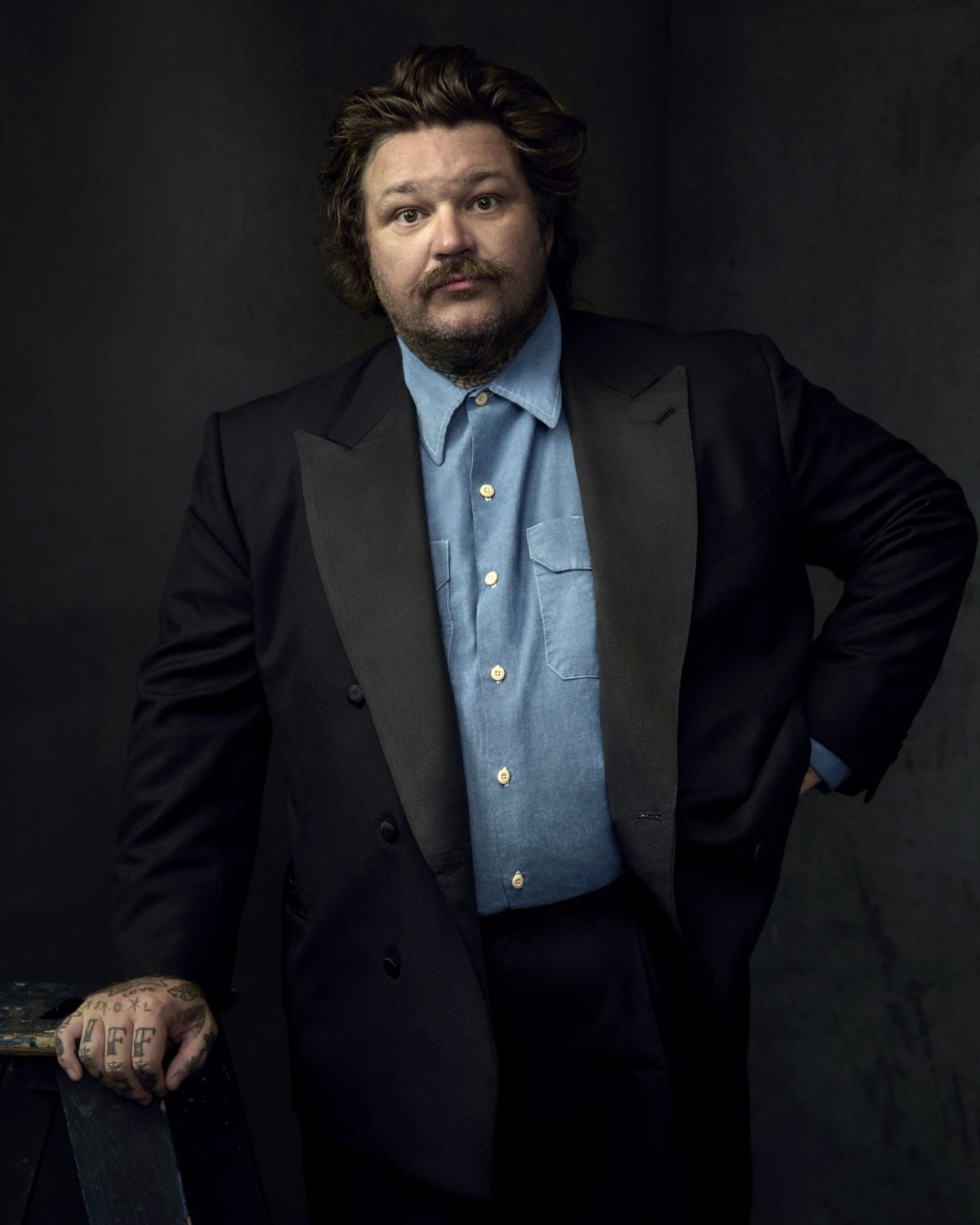 Portrait of a male in a black suit jacket and light blue button-down shirt, posing against a dark gray backdrop in a studio setting.
