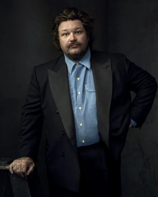 Portrait of a male in a black suit jacket and light blue button-down shirt, posing against a dark gray backdrop in a studio setting.