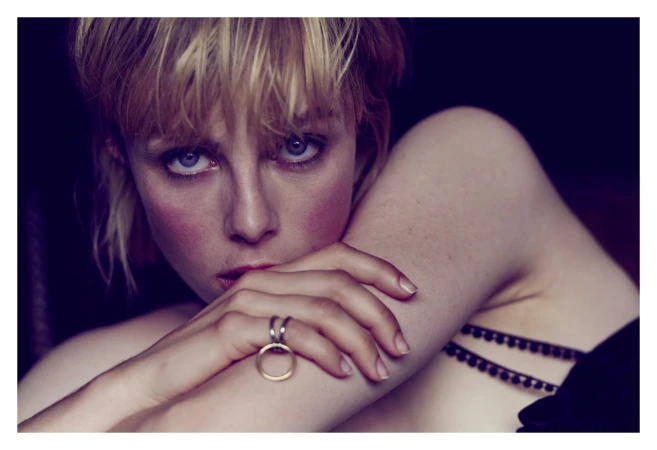 Close-up portrait of a woman with short blonde hair and blue eyes, resting her chin on her hand wearing rings.