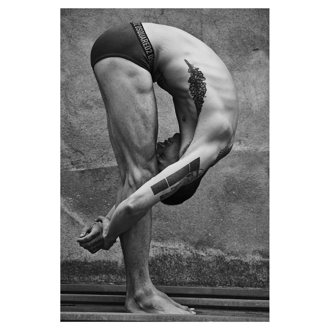 Black and white portrait of a muscular adult male performing a forward bend, touching his toes, showcasing tattoos against a concrete backdrop.