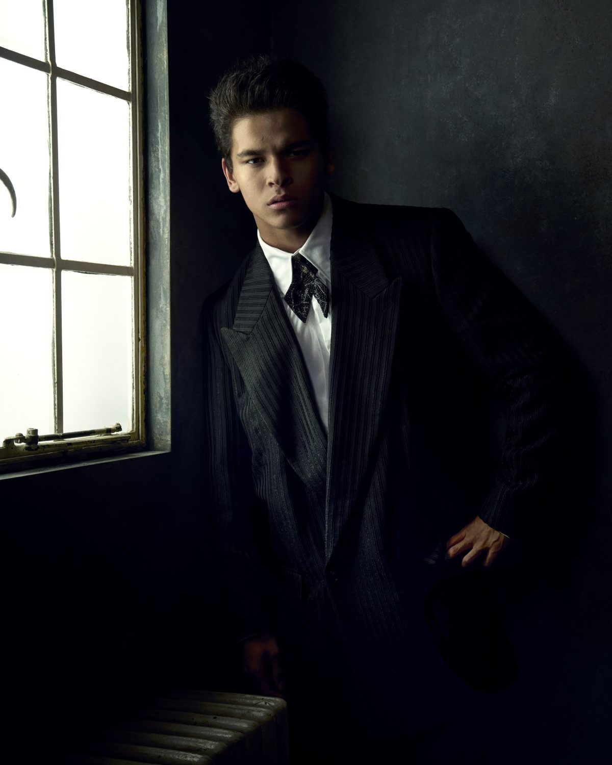 Portrait of a young adult male leaning against a window, wearing a dark pinstripe suit and bow tie in a dimly lit indoor setting.