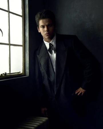 Portrait of a young adult male leaning against a window, wearing a dark pinstripe suit and bow tie in a dimly lit indoor setting.