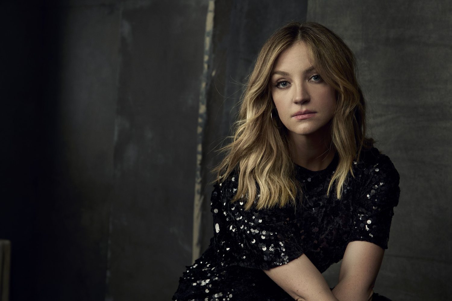 Portrait of a blonde woman with fair skin wearing a black sequined top, posing in an indoor studio against a dark grey textured backdrop.