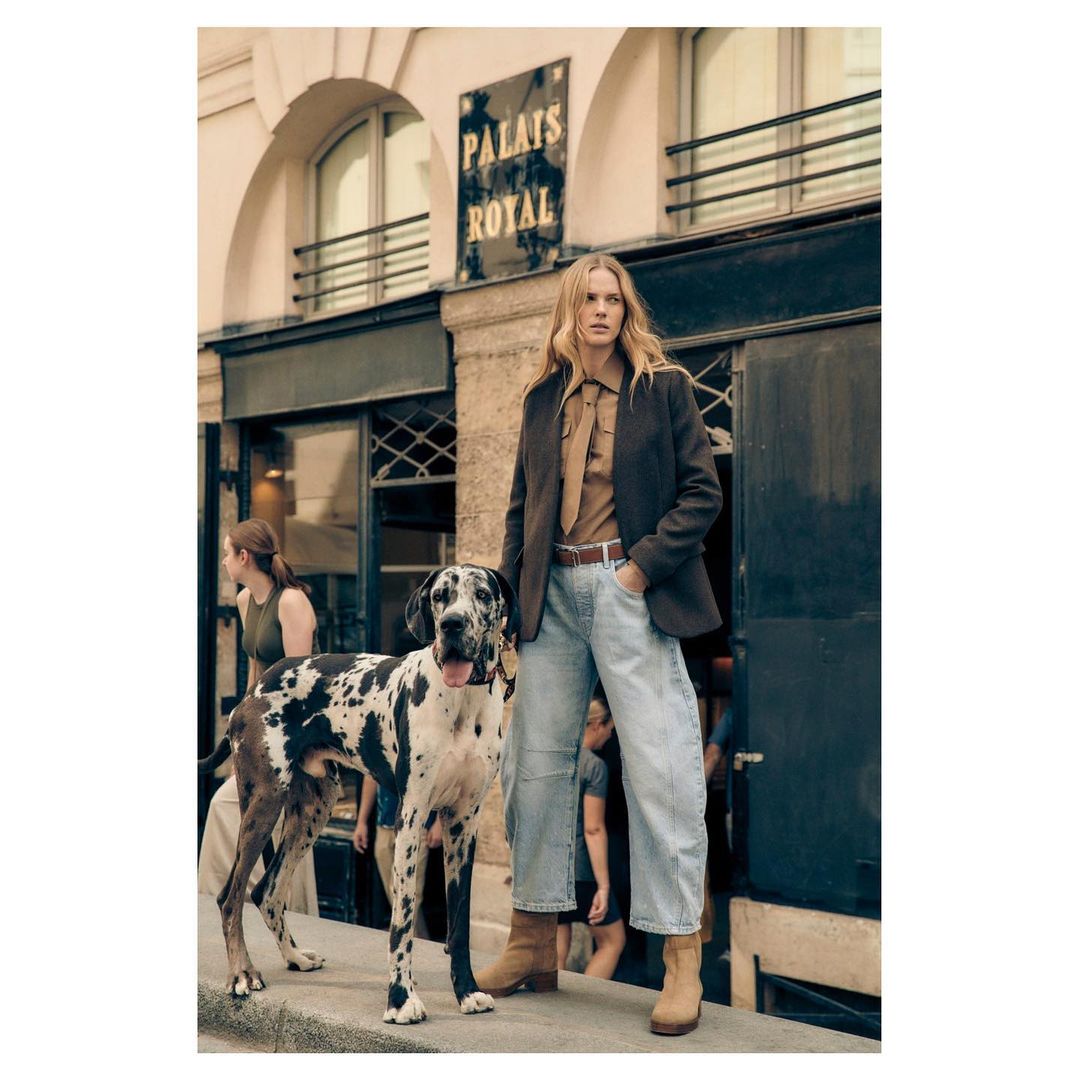 Blonde model poses with Great Dane dog on street in brown jacket, blue jeans and boots in front of building with 'Palais Royal' sign.