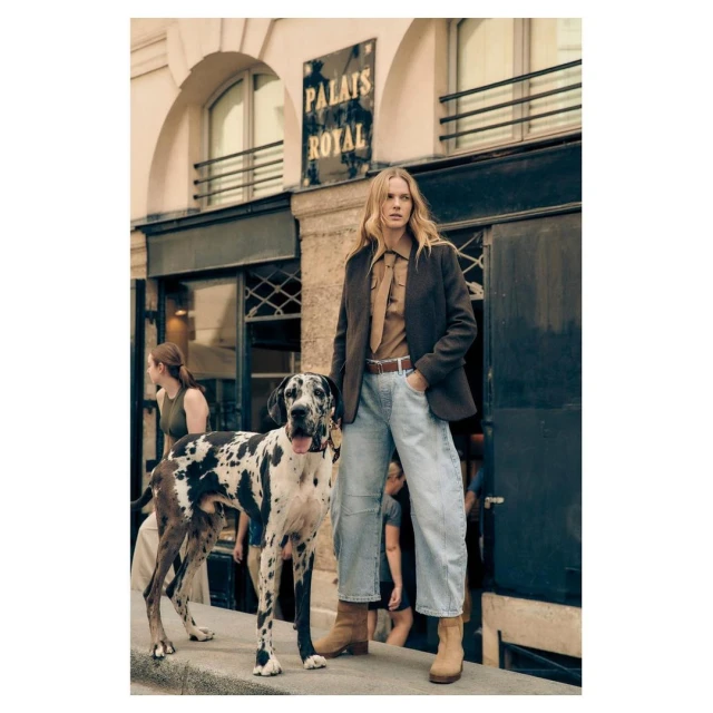 Blonde model poses with Great Dane dog on street in brown jacket, blue jeans and boots in front of building with 'Palais Royal' sign.