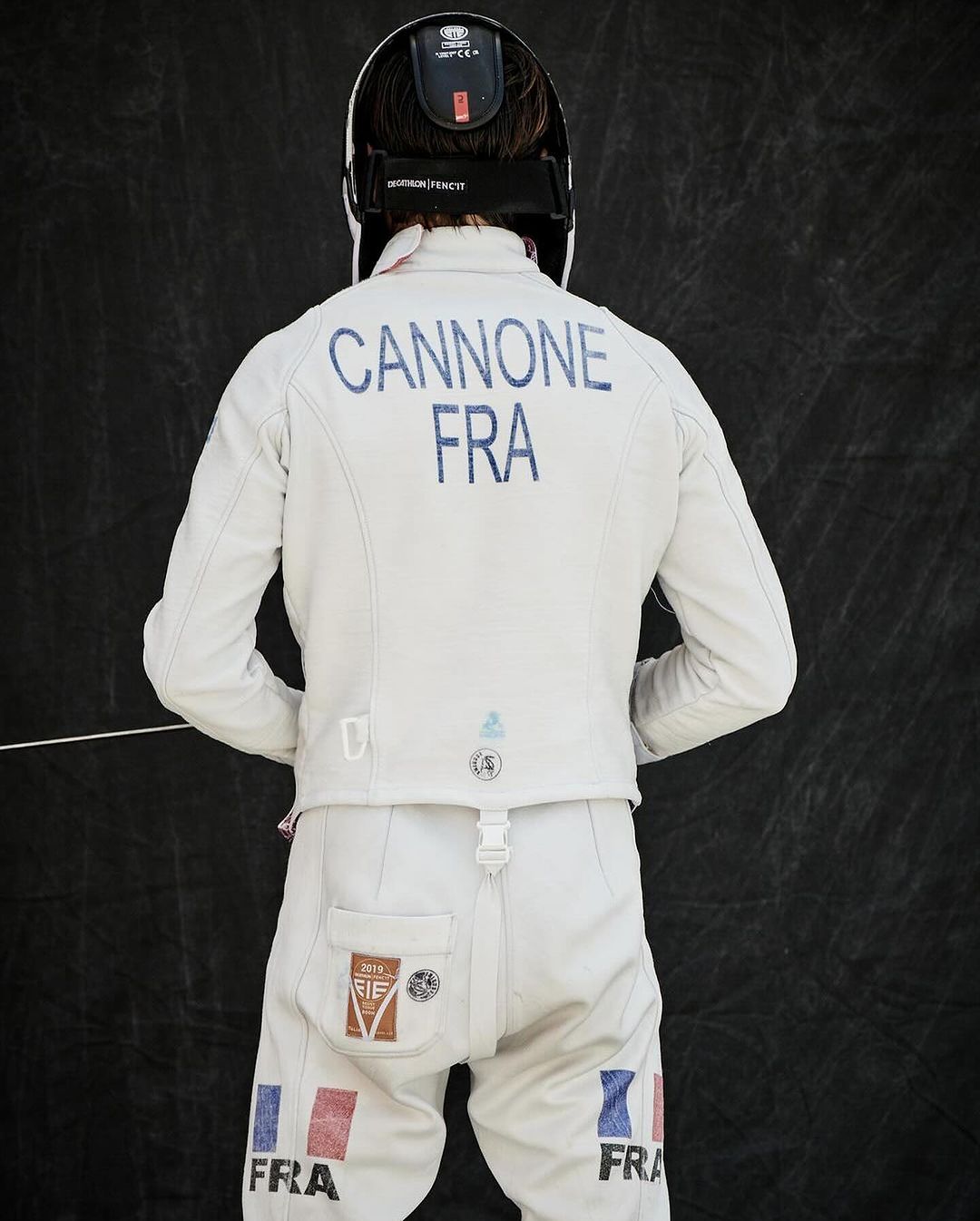 Back view of a fencer wearing a white uniform with "CANNONE FRA" printed on the back and a fencing mask against a dark background.