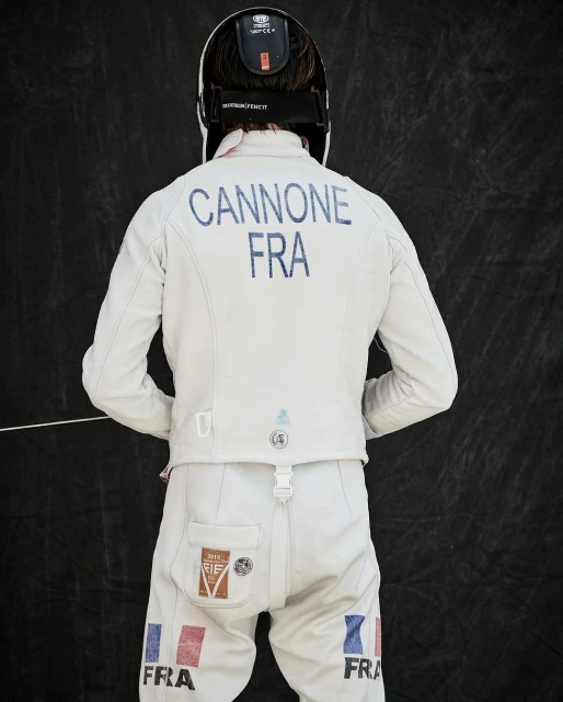 Back view of a fencer wearing a white uniform with "CANNONE FRA" printed on the back and a fencing mask against a dark background.