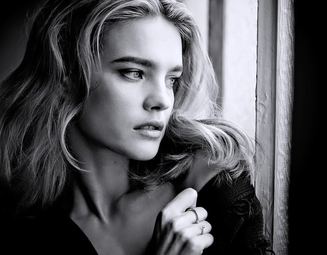 Black and white portrait of a woman with wavy blonde hair looking towards the light from a window, wearing a dark garment.