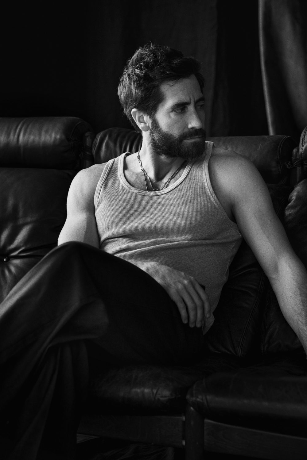 Black and white portrait of a bearded adult man wearing a tank top, seated on a black leather couch in an indoor setting.