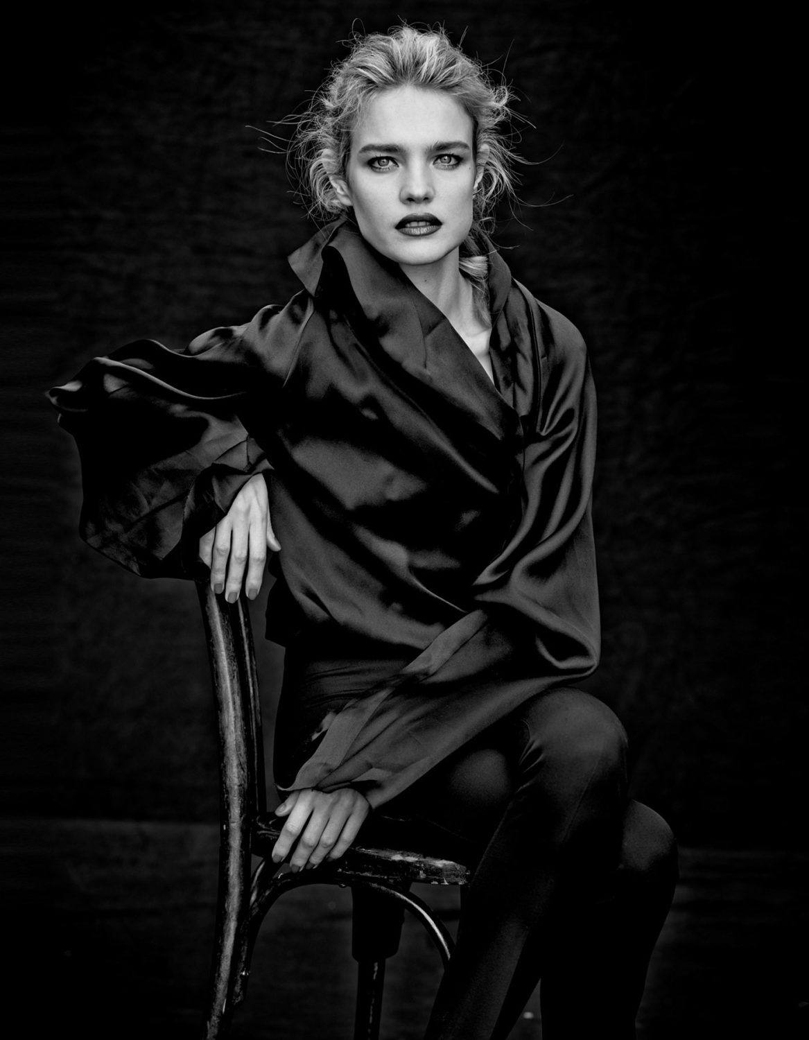Black and white portrait of a woman sitting on a chair, wearing a silk top and pants, with a dark background and soft lighting.