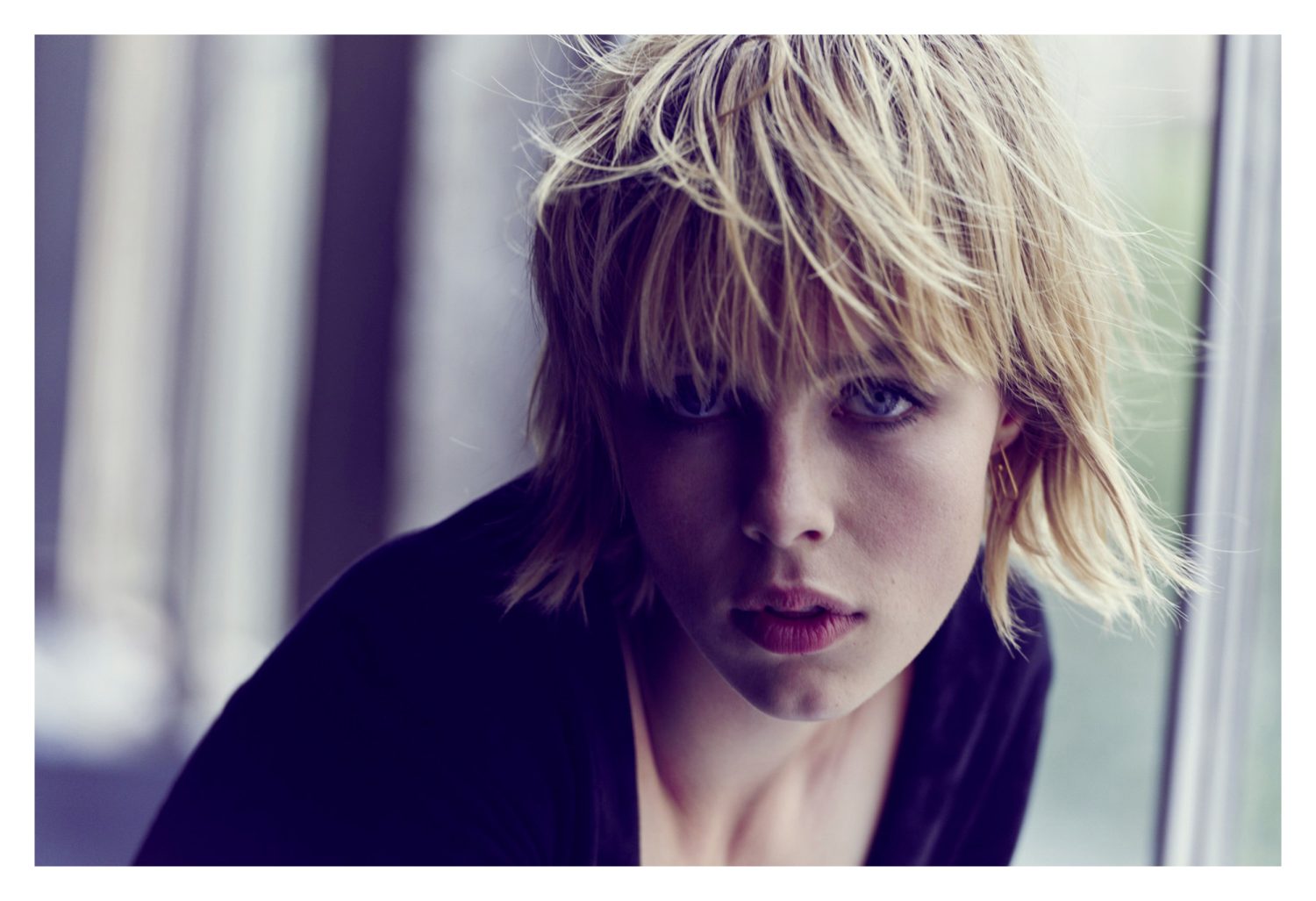 Close-up portrait of a woman with tousled blonde hair and bangs wearing a dark top, directed by Matthew Brookes.