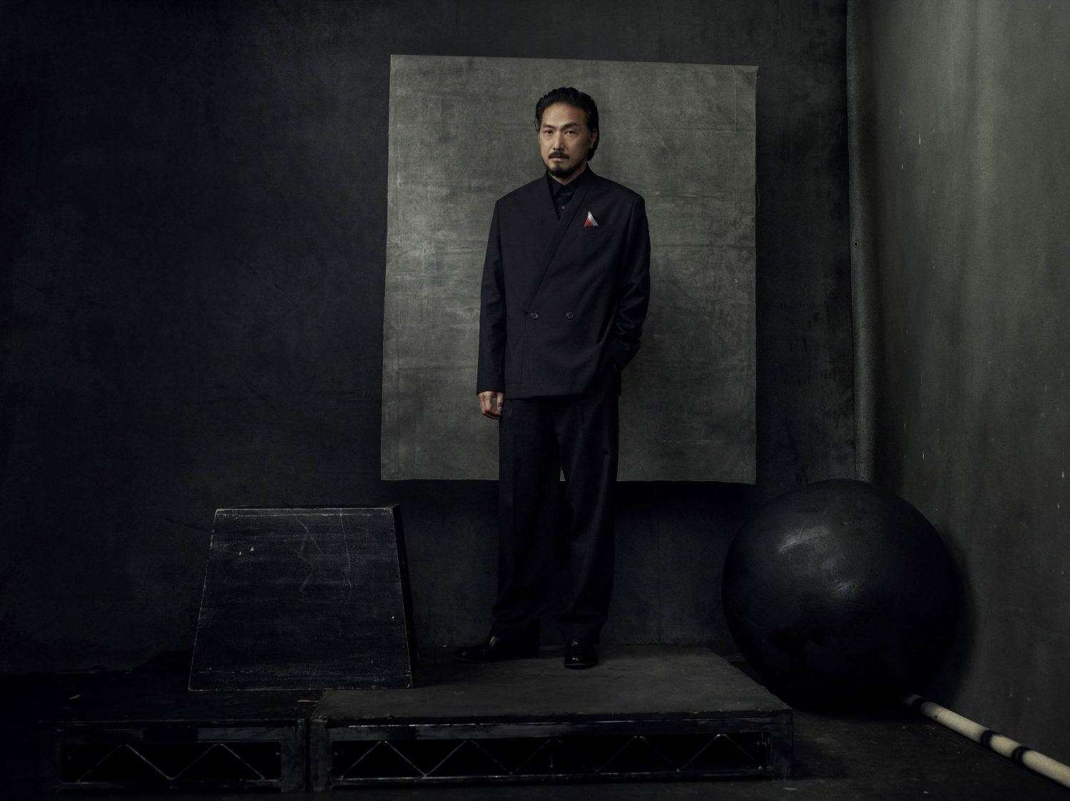 Portrait of an adult man wearing a black suit, standing in front of a mottled gray backdrop in a dark, low-light studio setting.