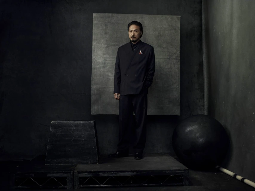Portrait of an adult man wearing a black suit, standing in front of a mottled gray backdrop in a dark, low-light studio setting.