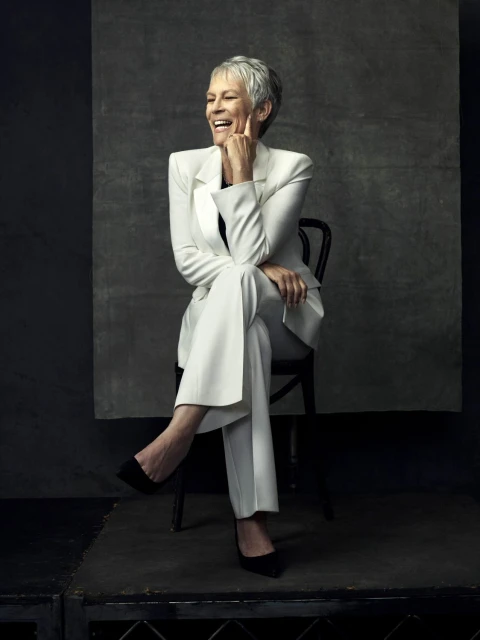 Jamie Lee Curtis sits on a black chair wearing a white suit in a studio setting with a grey mottled background, looking to the side.