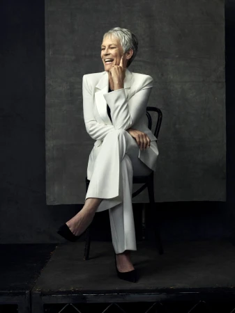 Jamie Lee Curtis sits on a black chair wearing a white suit in a studio setting with a grey mottled background, looking to the side.