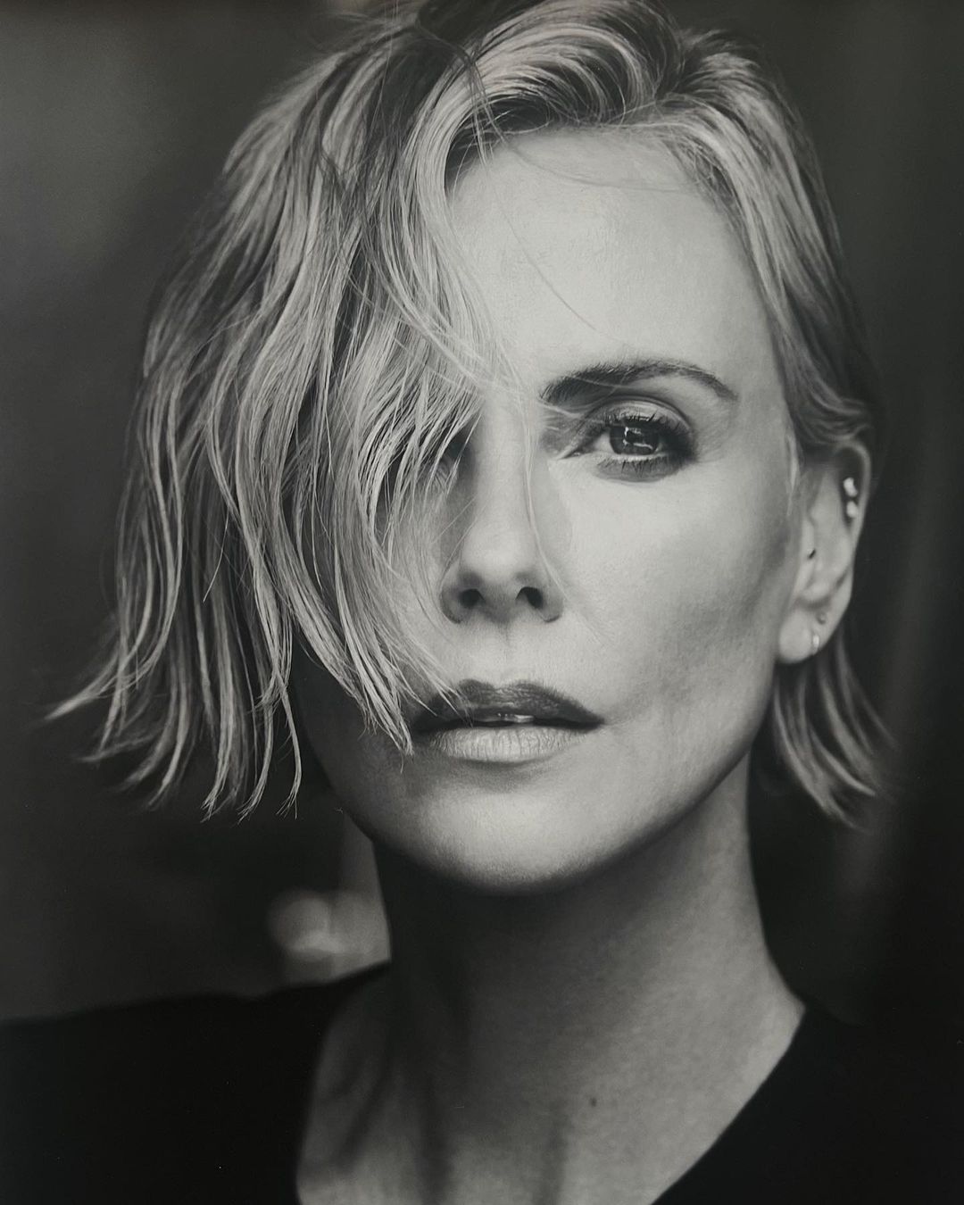 Monochrome portrait of adult woman with short, layered blonde hair, fair skin, and dark eyes, in a close-up headshot against a dark backdrop.