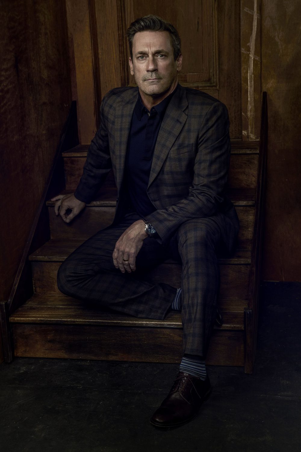Adult male in a grey plaid suit sitting on wooden stairs, wearing a dark shirt and brown leather shoes, indoors with soft lighting.