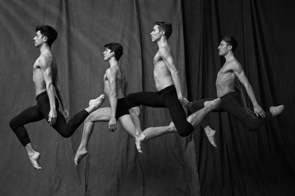 Four male ballet dancers in mid-leap against a gray backdrop, captured in a monochrome studio shot.