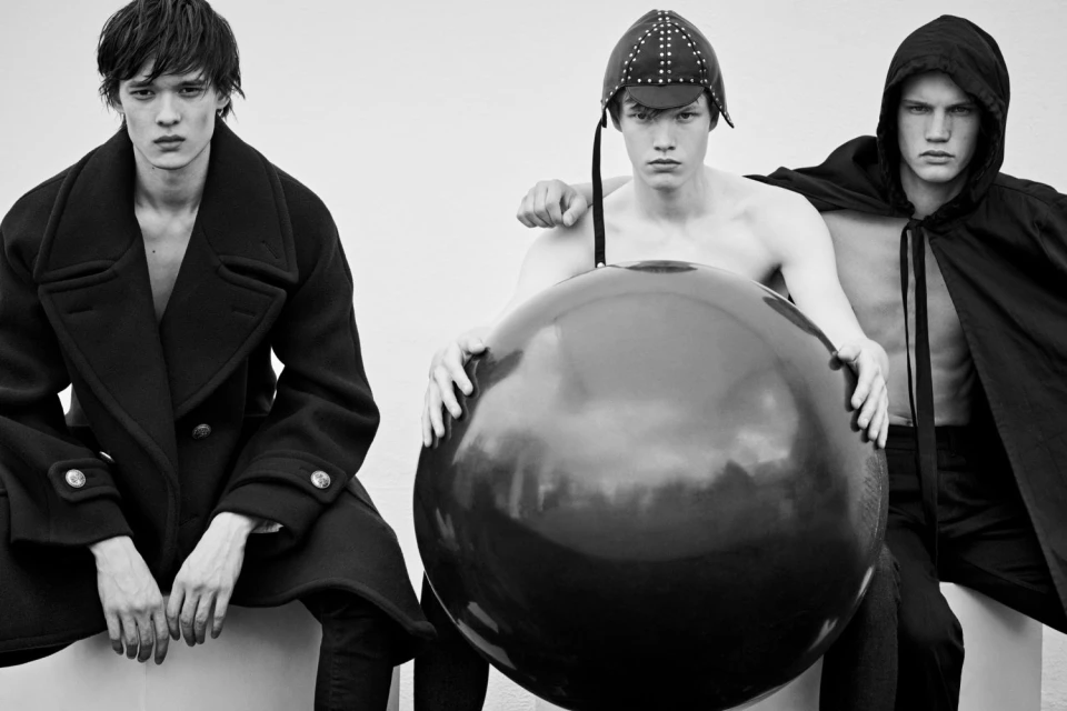 Three fair-skinned adult male models pose in black and white, seated on a white cube with a neutral backdrop, with one holding a black sphere.