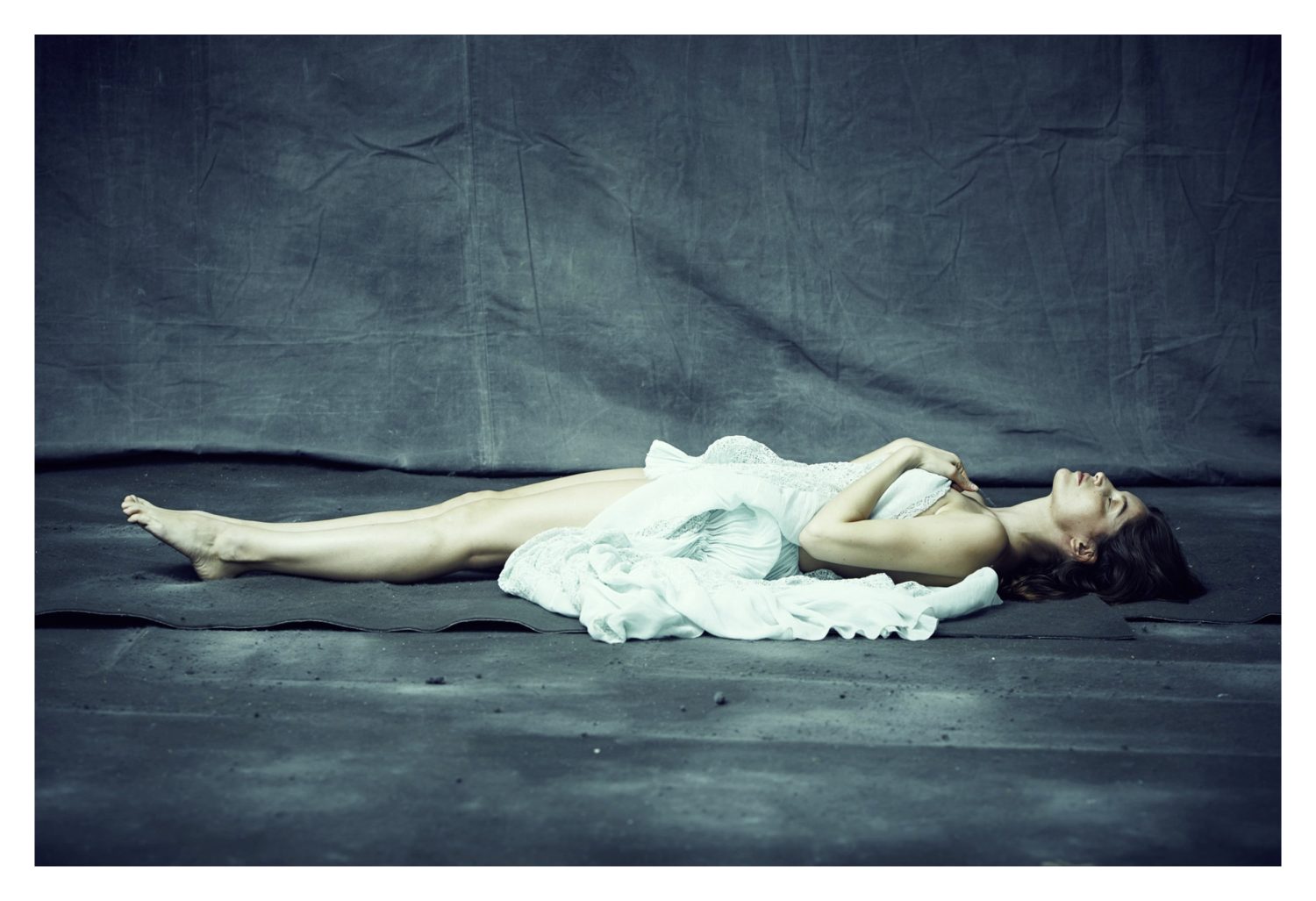 Elegant woman lying down on a gray surface, draped in a flowing white fabric in a full shot against a draped studio backdrop.