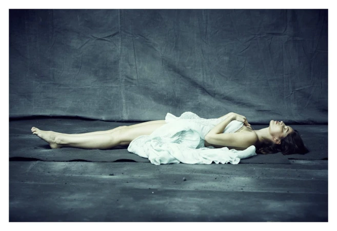 Elegant woman lying down on a gray surface, draped in a flowing white fabric in a full shot against a draped studio backdrop.