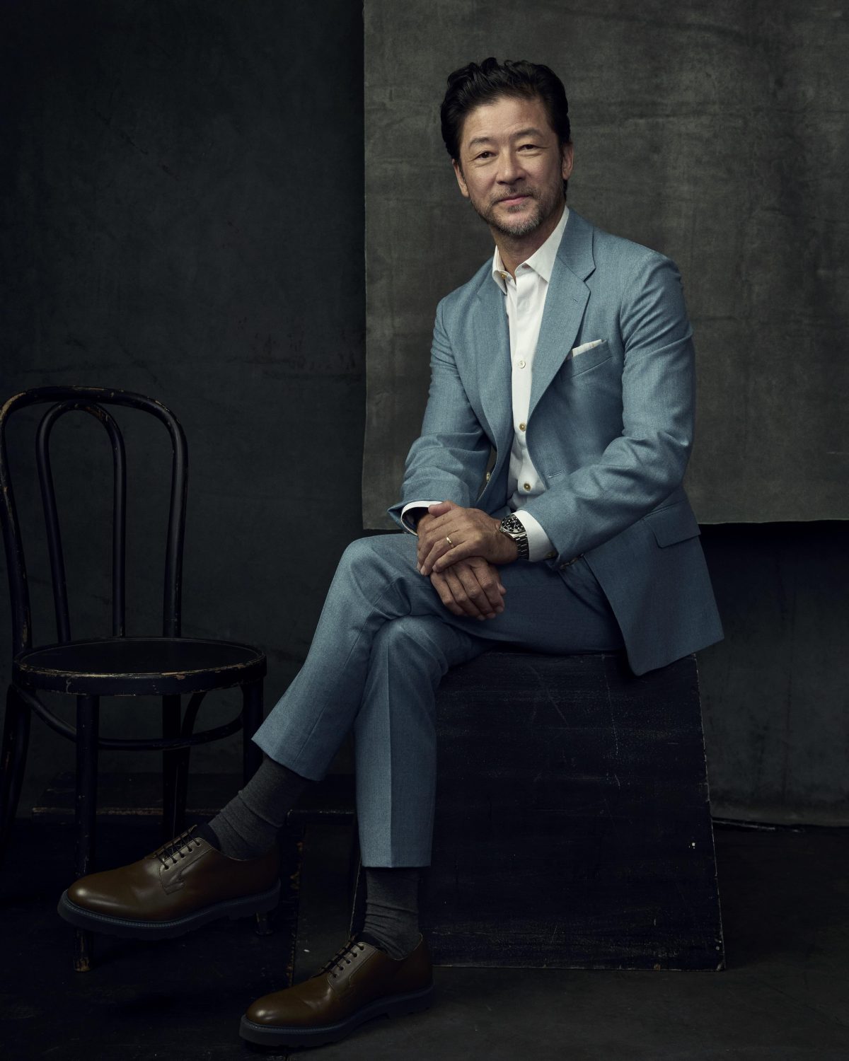 Middle-aged man in a light blue suit seated on a dark box in a studio setting by Matthew Brookes