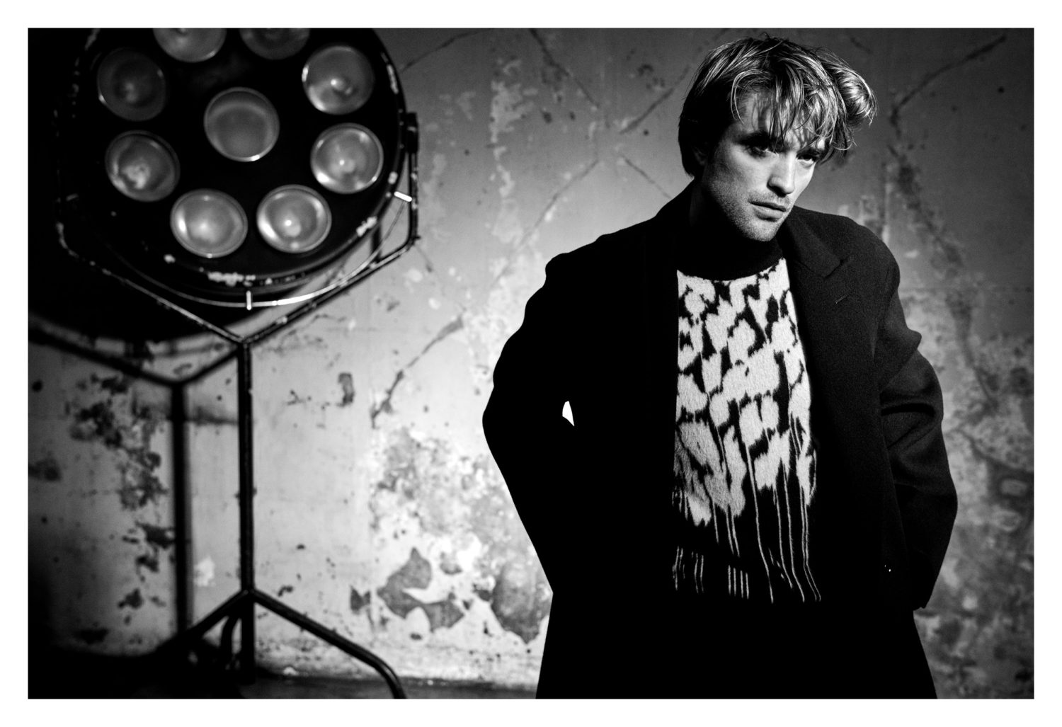 Monochrome portrait of adult male model wearing patterned sweater and dark coat in front of light fixture and distressed wall.
