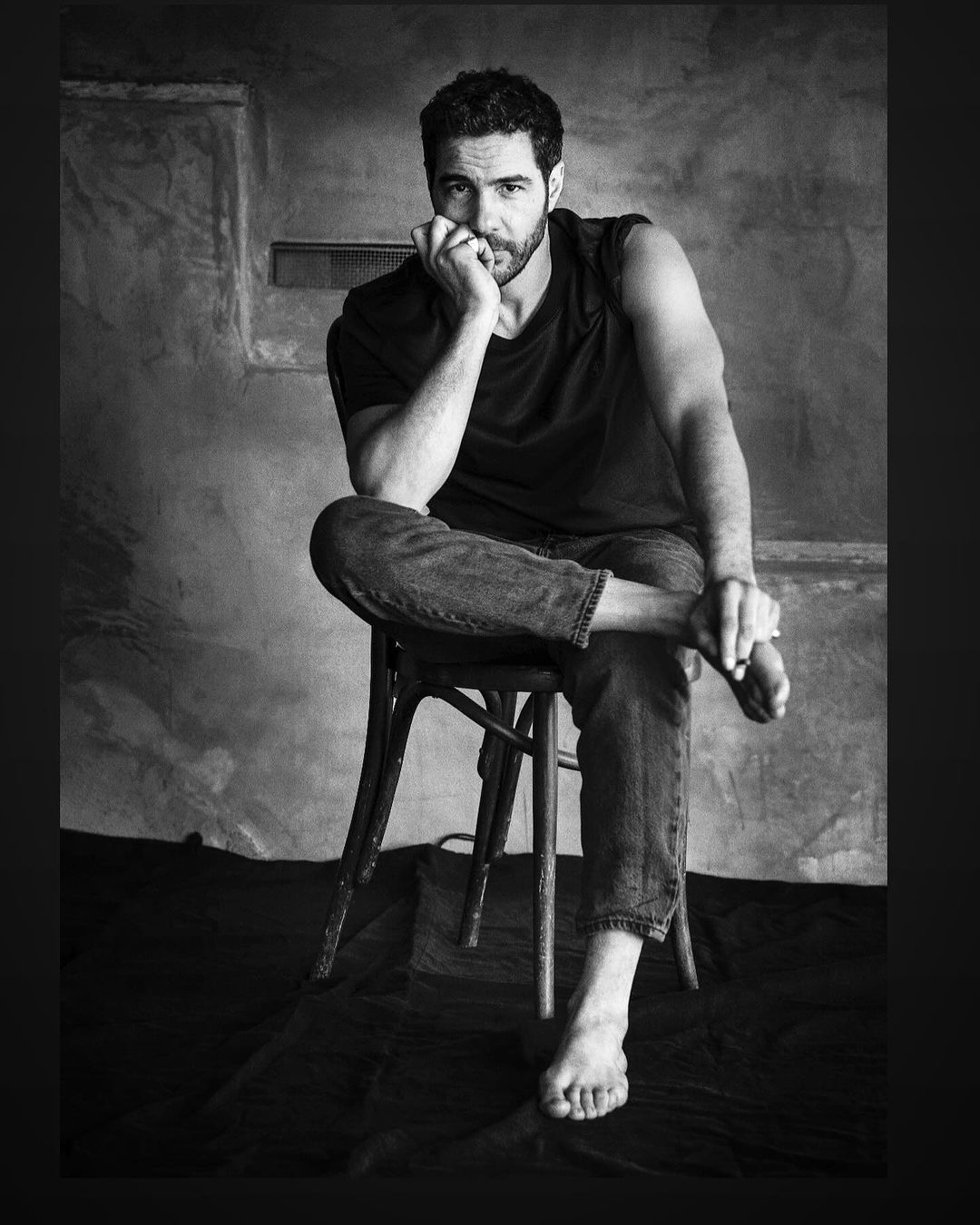 Monochrome portrait of a man sitting barefoot on a wooden chair against a textured concrete wall