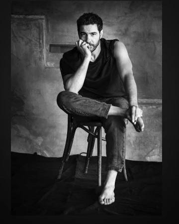 Monochrome portrait of a man sitting barefoot on a wooden chair against a textured concrete wall