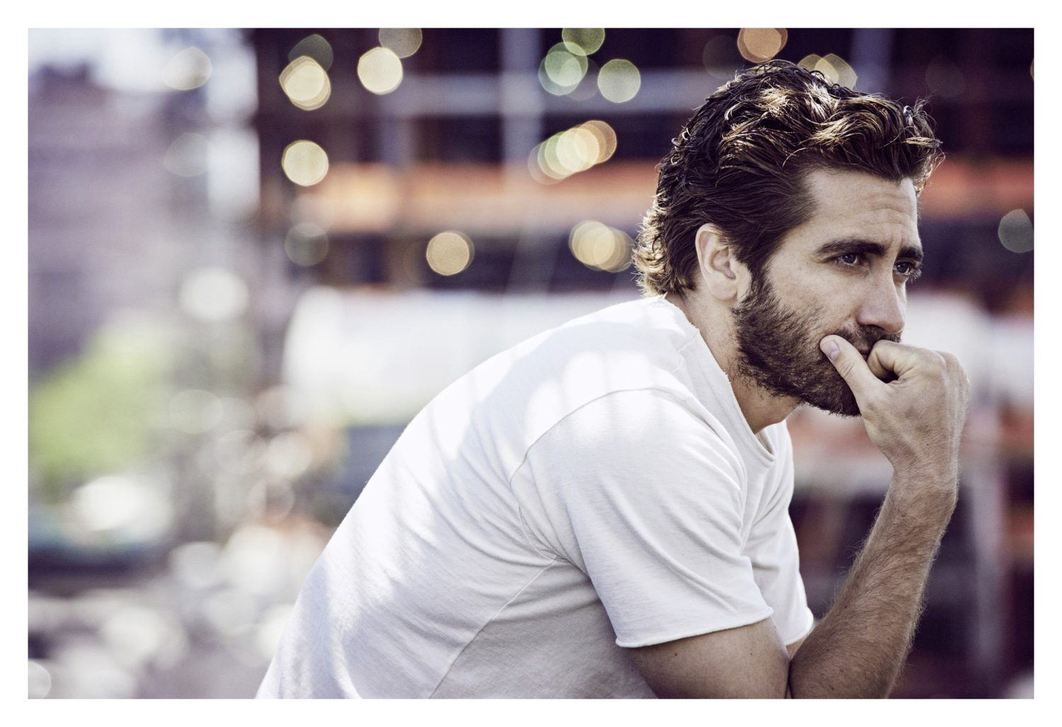 Portrait of a bearded adult male in a white t-shirt, gazing to the side with a thoughtful expression in a blurry, urban setting.