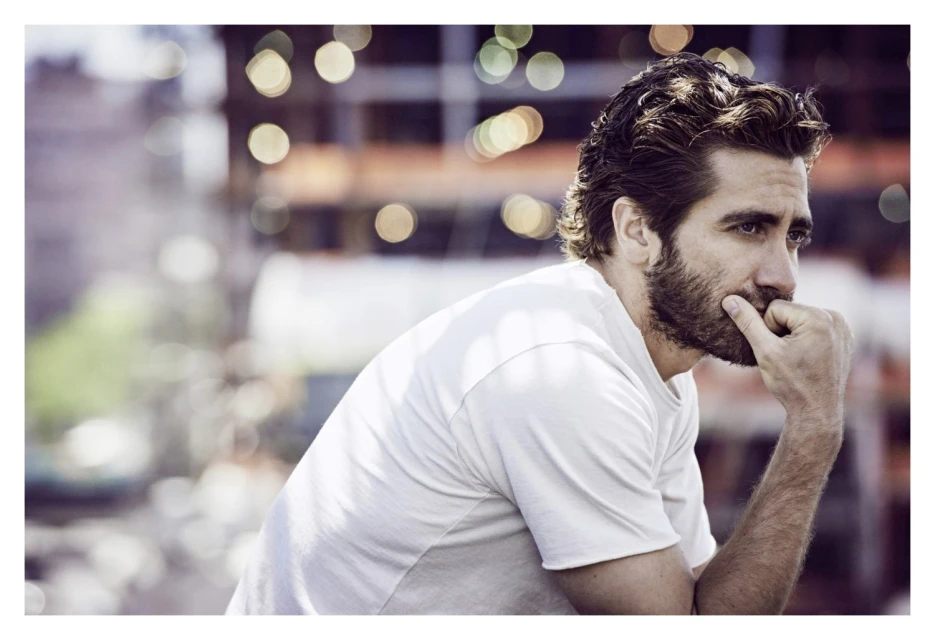 Portrait of a bearded adult male in a white t-shirt, gazing to the side with a thoughtful expression in a blurry, urban setting.