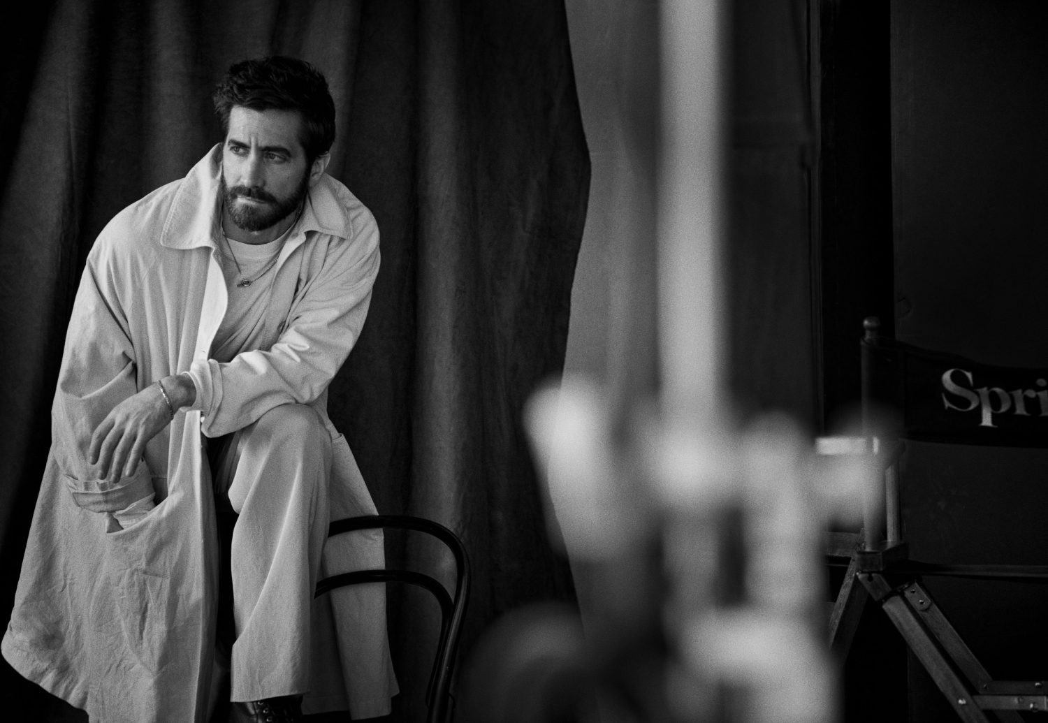 Black and white portrait of bearded man in an overcoat sitting on a chair against draped backdrop looking left with a serious expression.