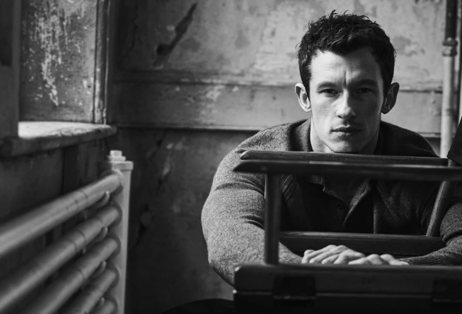 Black and white portrait of a handsome man with short dark hair, wearing a sweater, leaning against a chair, in a weathered interior.