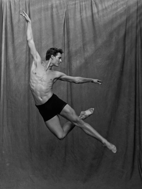 Black and white shot of a shirtless male ballet dancer leaping mid-air against a draped backdrop, showcasing athleticism and artistry.