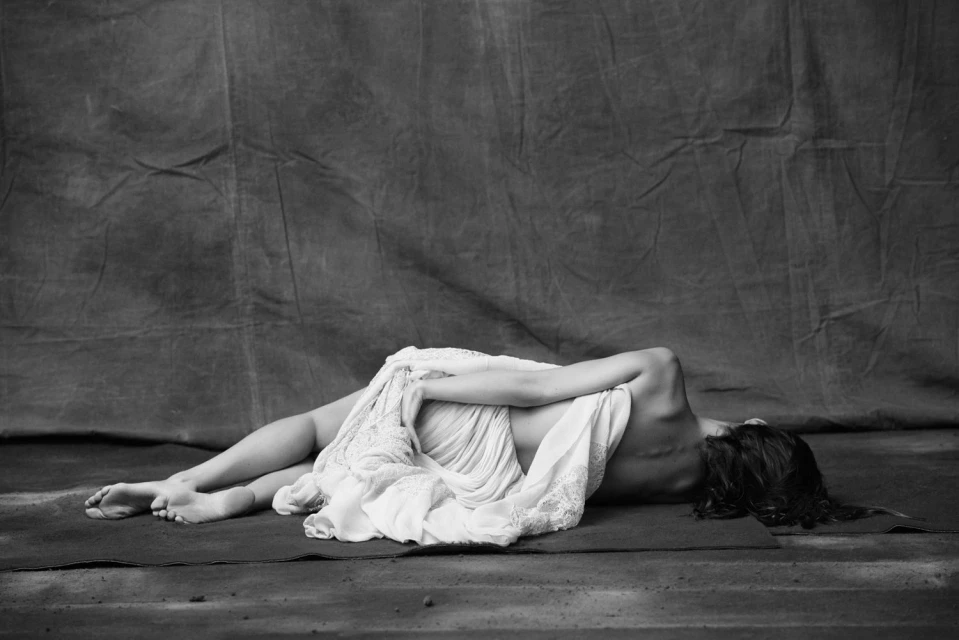 Black and white portrait of a woman lying down and partially covered with a white fabric against a textured grey backdrop.