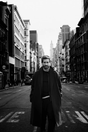 Black and white portrait of actor Jake Gyllenhaal walking down a city street wearing a long dark coat