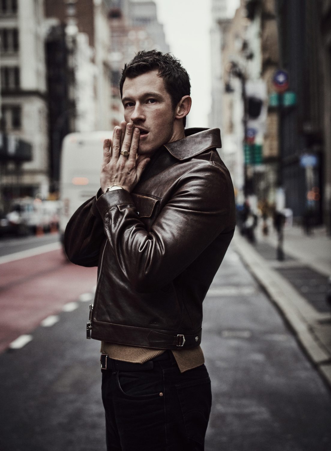 Man wearing a dark brown leather jacket posing on a city street holding hands to his face, directed by Matthew Brookes.